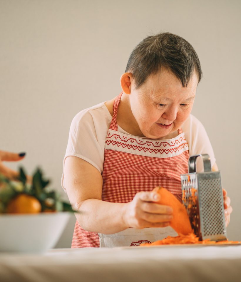 elderly woman with Down syndrome gratefully learns to grate carrots with the guidance of a teacher
