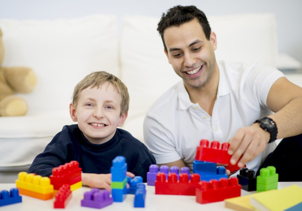 A caregiver is playing with a boy that has a physical disability. They are playing with plastic blocks together.